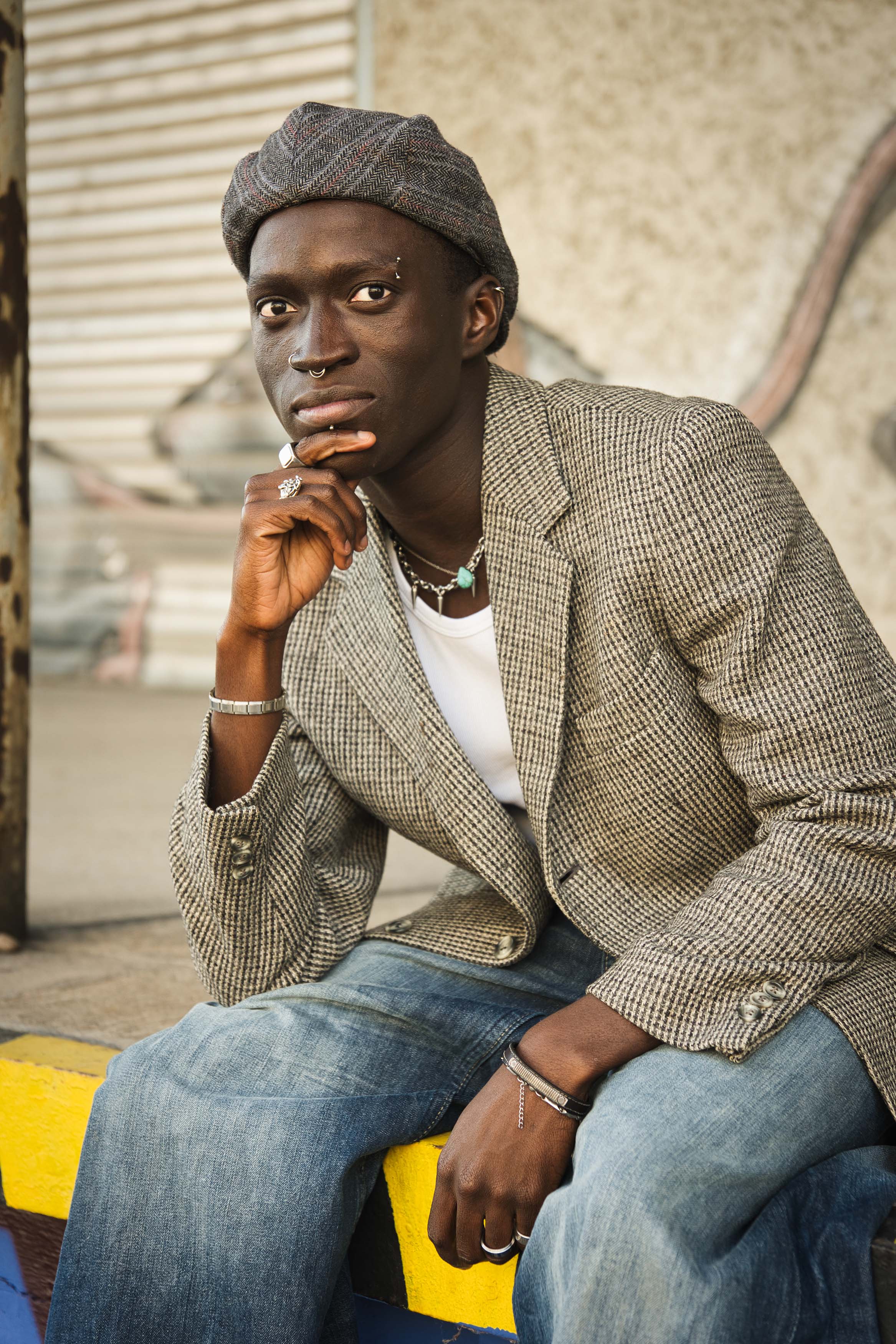 Fashion Porträt von Kelvin Addai mit Barett und karierter Jacke, sitzend in urbaner Umgebung in Linz, im Streetstyle-Look