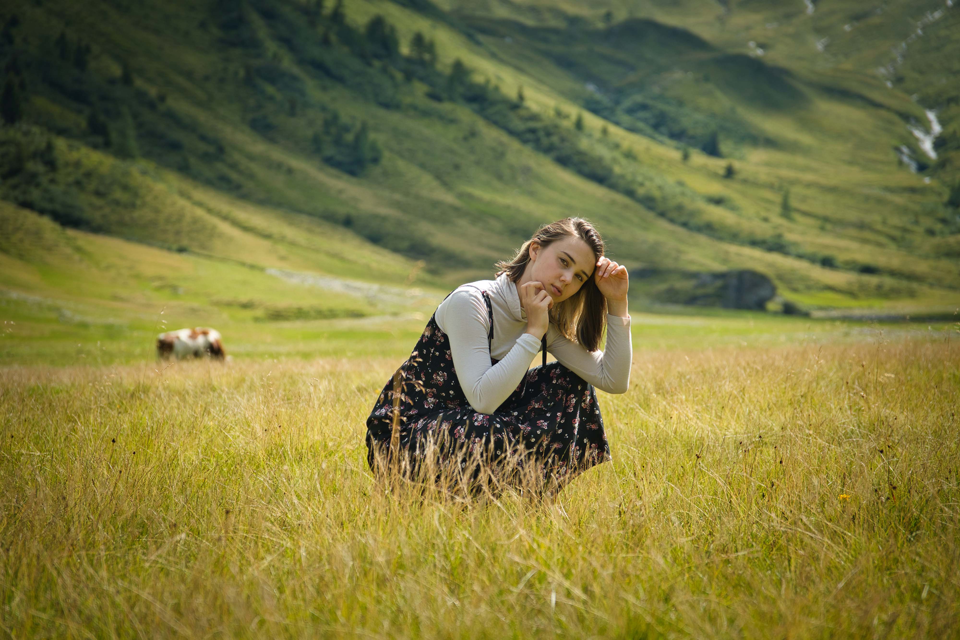 junge Frau hockt nachdenklich auf einer Bergwiese in den Alpen, im Hintergrund grüne Hügel und eine Kuh auf der Alm
