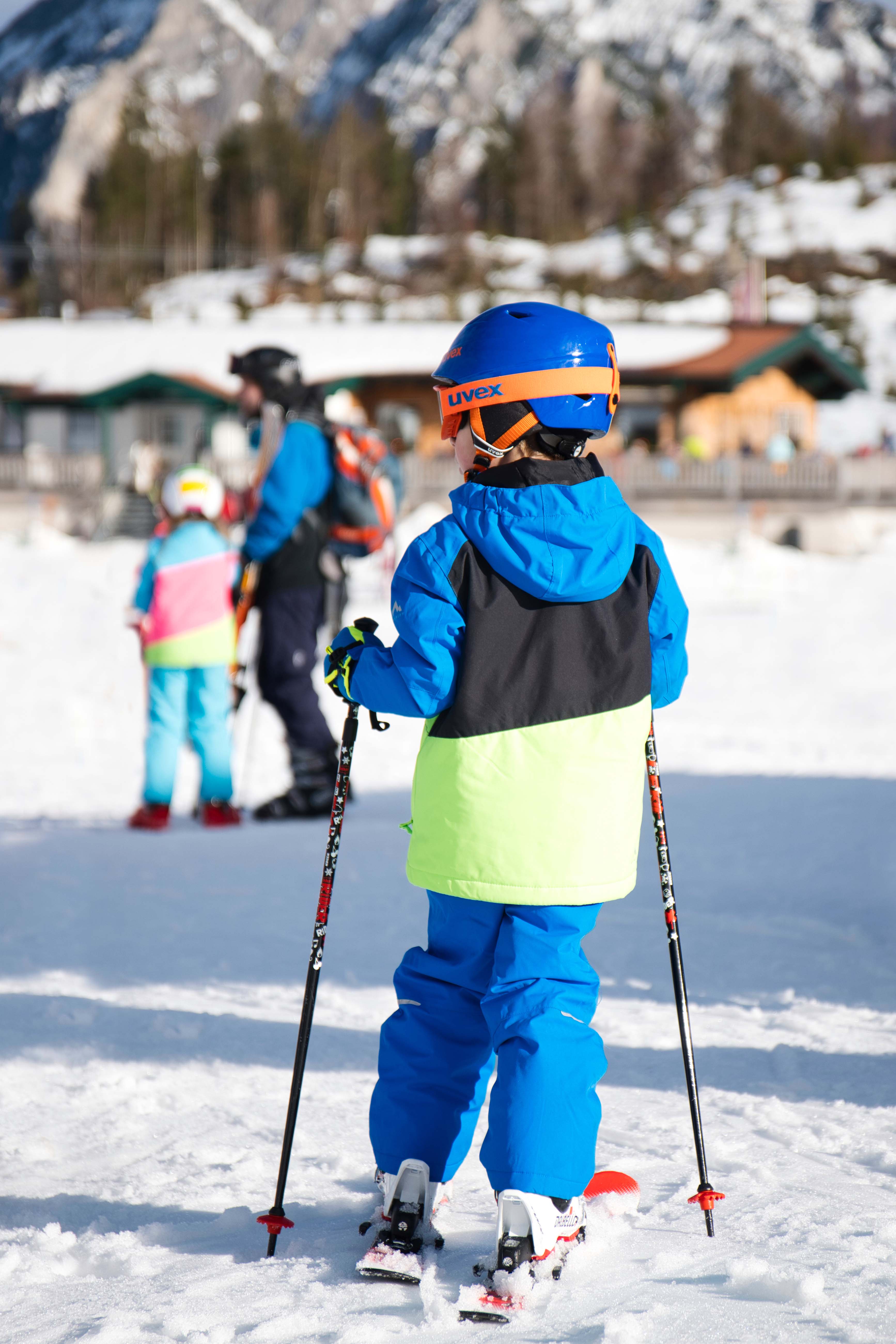 Kind in blauer Skibekleidung mit Skistöcken im Postalm Winterpark, weitere Skifahrer und Hütten im Hintergrund