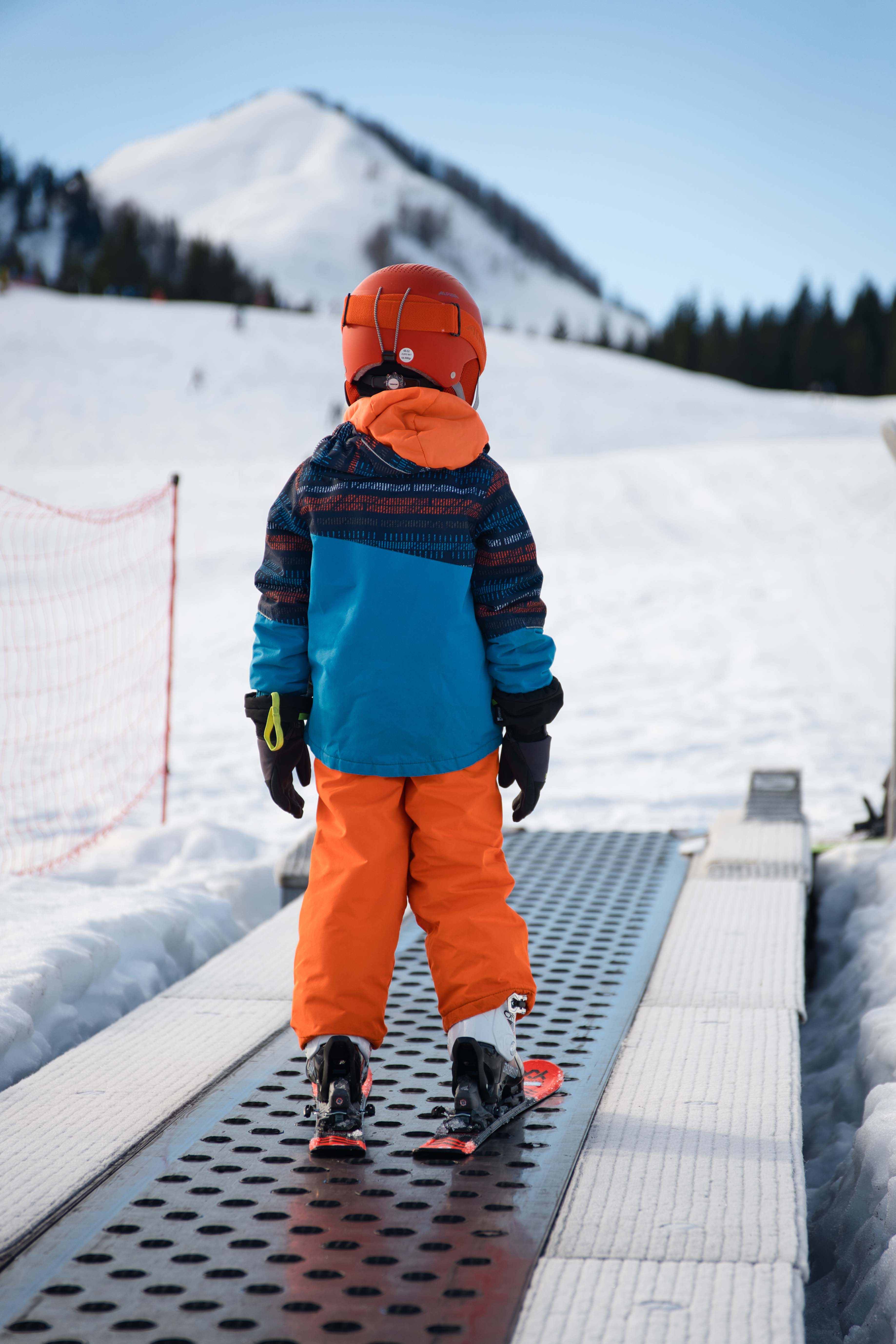 Kind in bunter Skibekleidung auf Förderband im Postalm Winterpark, verschneiter Berg im Hintergrund